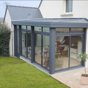 Véranda en aluminium gris anthracite avec toit plat, grandes baies vitrées coulissantes. Extension lumineuse donnant sur le jardin et la terrasse. Sapin de Noël visible.