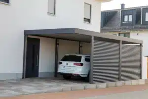 Carport adossé gris foncé devant une maison moderne blanche. Un break VW y est garé, protégé par une paroi latérale à lattes horizontales.