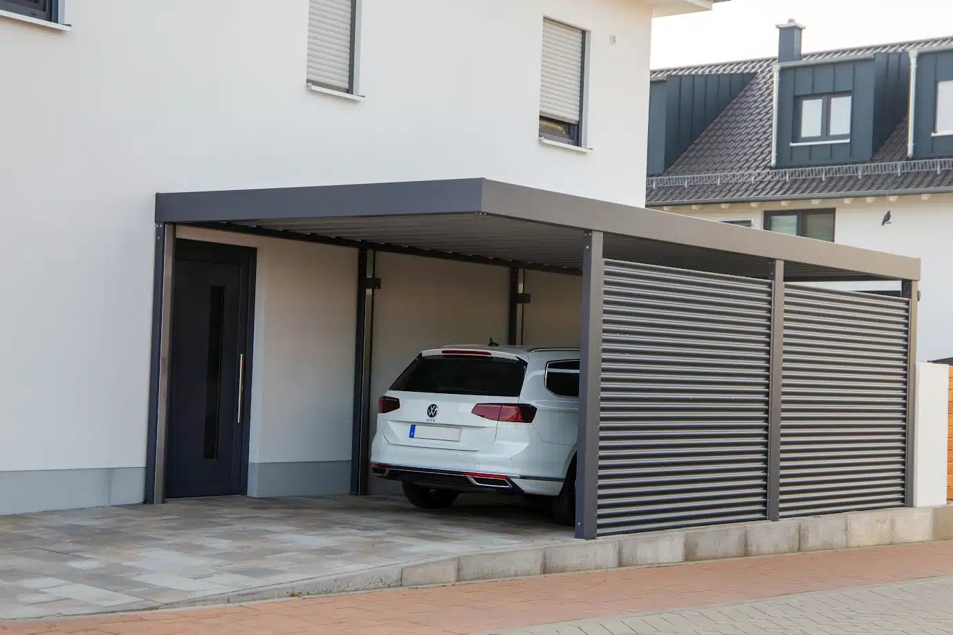 Carport adossé gris foncé devant une maison moderne blanche. Un break VW y est garé, protégé par une paroi latérale à lattes horizontales.