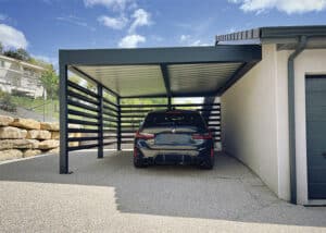 Abri voiture moderne en aluminium gris, avec lattes décoratives, couvrant une BMW. Allée en moquette de pierre près d'une maison et de rochers.