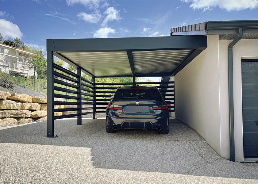 Abri voiture moderne en aluminium gris, avec lattes décoratives, couvrant une BMW. Allée en moquette de pierre près d'une maison et de rochers.